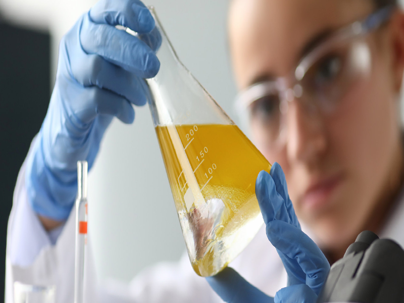 Científica de laboratorio con guantes y gafas, examinando líquido amarillo en matraz Erlenmeyer.